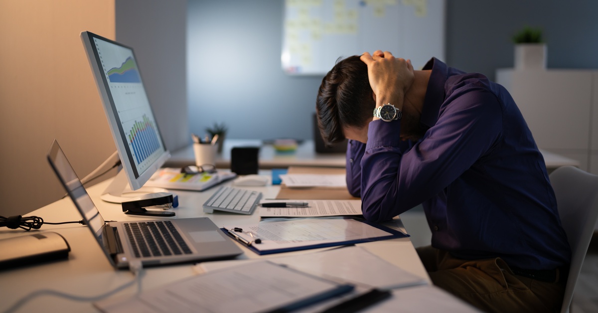 A man sits at a desk with a laptop, monitor, and a lot of paperwork. He has his head down with his hands behind his neck.