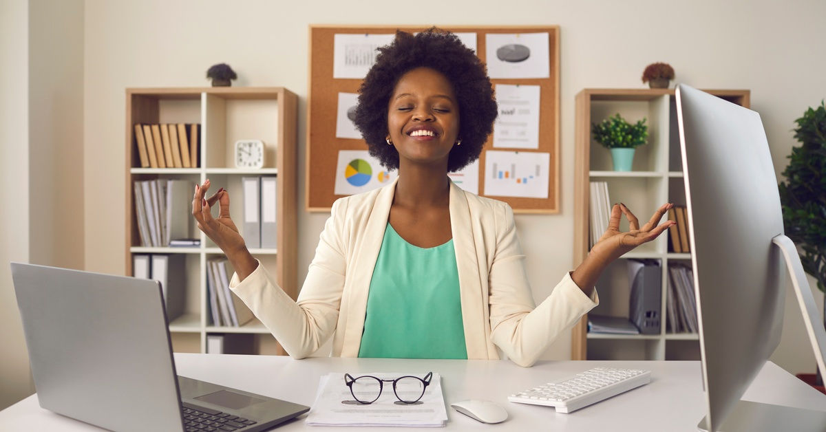A young Black businesswoman meditates at her desk with her hands raised in a mudra gesture, embracing calm at work.